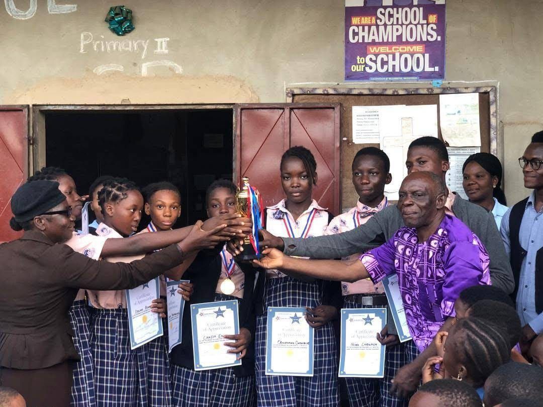 Des élèves tiennent des certificats encadrés et un trophée aux côtés d’adultes, devant une porte marquée Primary II.