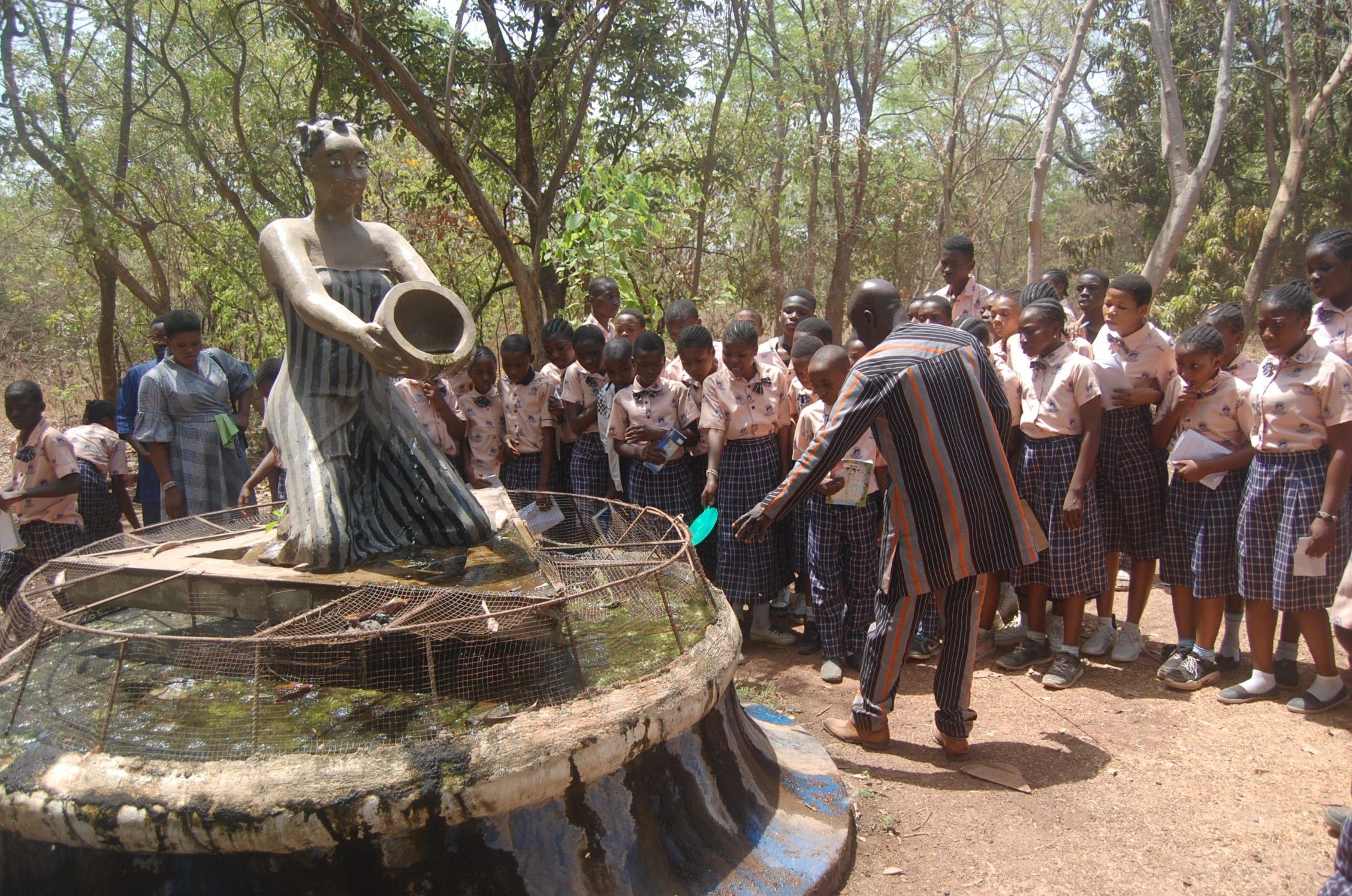 Des élèves en uniforme à carreaux entourent un bassin de pierre et une sculpture pendant qu’un adulte leur montre l’eau.